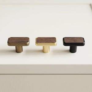 May include: Three cabinet knobs with rectangular wooden tops. The knobs have different metal finishes: brass, gold, and black. The knobs are designed for furniture and home decor.