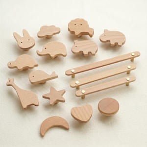 Op de afbeelding: Een verzameling houten kastknoppen en -grepen in verschillende vormen, waaronder een konijn, een wolk, een leeuw, een nijlpaard, een schildpad, een walvis, een giraffe, een ster, een halve maan, een hart en een ronde knop. De grepen zijn rechthoekig met messing accenten.