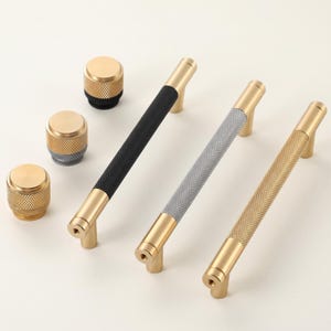 May include: A collection of cabinet hardware, including three textured bar pulls in black, gray, and gold, and three matching round knobs. The pulls have gold-toned ends and a textured center. The knobs are gold with black or gray accents.