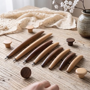 Puede incluir: Una colección de ganchos de ganchillo y pomos de madera en varios tonos de marrón. Los ganchos están dispuestos en diagonal, mostrando su diseño cónico. También se muestran varios pomos redondos, junto a un jarrón con flores blancas.