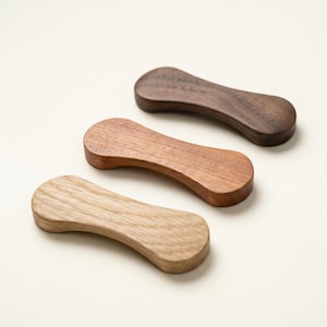 May include: Three wooden chopstick rests in varying shades of brown. The rests are curved with a rounded hourglass shape. The top rest is dark brown, the middle is medium brown, and the bottom is light brown.