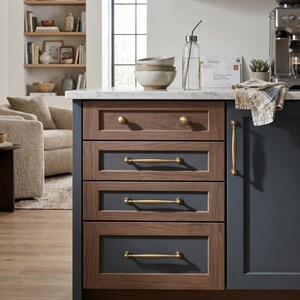 May include: A kitchen island featuring dark wood drawers with brass handles. The drawers are framed in medium brown wood with grey-blue centre panels. A clear glass water bottle and bowls are on the countertop.