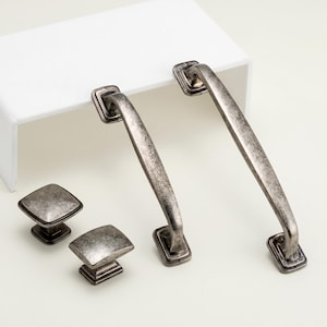 Può includere: Un set di ferramenta per mobili in stile antico, tra cui due maniglie e due pomelli. La ferramenta ha una finitura argentata anticata con piastre quadrate e una superficie strutturata. Le maniglie sono lunghe circa 15 cm.