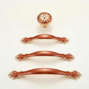 May include: A set of decorative cabinet hardware featuring a round knob and three matching pull handles. The knob and handles have a brown finish with gold-colored accents. The hardware is designed with an ornate, vintage style.