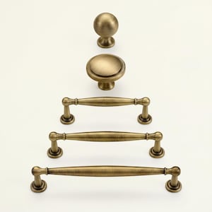 May include: An assortment of antique brass cabinet hardware, including a round knob, a flat-topped knob, and three different sizes of bar pulls. The hardware has a warm, aged finish, and is arranged vertically against a white background.