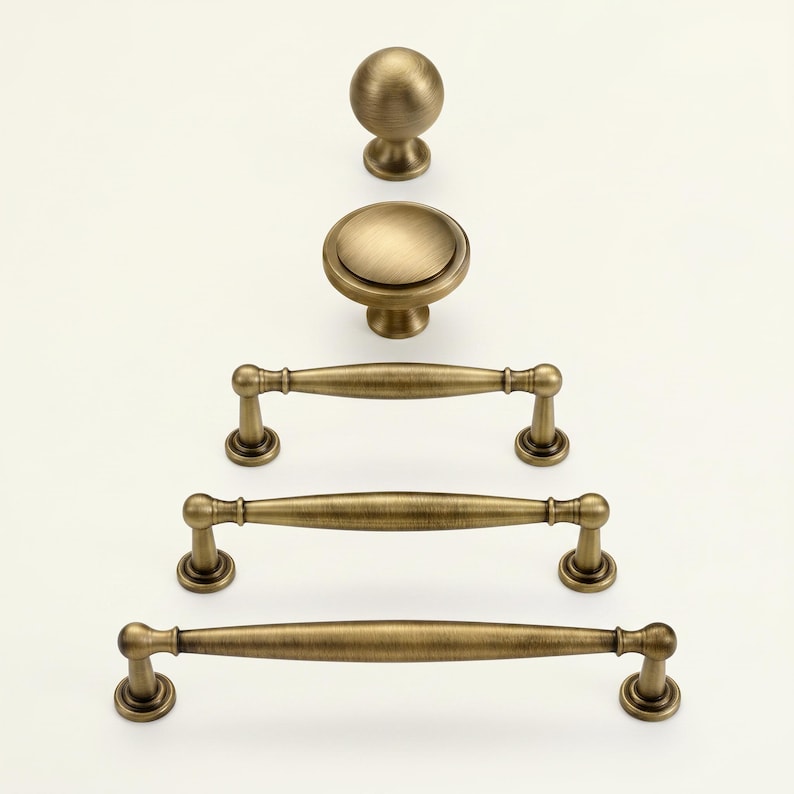 May include: A selection of antique brass cabinet hardware, featuring a round knob, a flat-topped knob, and three bar pulls of varying lengths. The hardware has a warm, aged finish, and is arranged vertically against a white backdrop.
