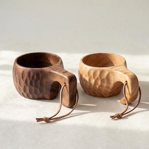 May include: Two wooden Kuksa cups with carved exteriors and attached leather straps. One cup is a dark brown, the other is a lighter tan color. The cups have a handle and are designed for outdoor use.
