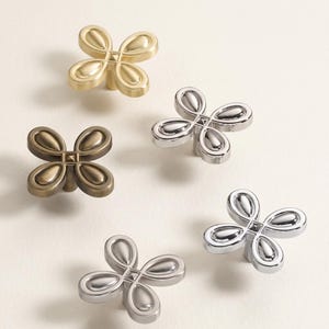 May include: Five decorative cabinet knobs in various metallic finishes. The knobs feature a four-petal design with rounded edges. Finishes include gold, bronze, and silver tones. The knobs are likely made of metal and are suitable for furniture or cabinetry.