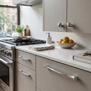 Puede incluir: Una cocina moderna con armarios de color gris claro y herrajes de acero inoxidable. Los armarios tienen pomos en forma de p&aacute;jaro y tiradores de caj&oacute;n curvos. Un cuenco de fruta c&iacute;trica se encuentra sobre la encimera.