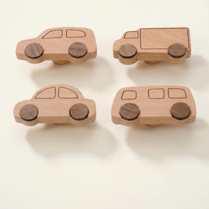 May include: Four wooden cabinet knobs in the shape of vehicles: a car, a van, a bus, and a truck. Each knob is light-colored with dark brown wheels. The knobs are designed with simple lines and shapes, suitable for a child's room or a playful decor.