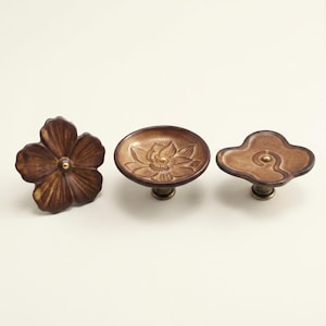 May include: Three decorative cabinet knobs in a warm brown hue. The knobs feature floral and cloud designs with brass accents. The flower knob has five petals, the lotus knob has a detailed flower, and the cloud knob has a swirl design.