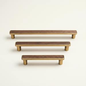May include: Three cabinet pulls with a rectangular walnut wood center and brass-colored metal frame and posts. The pulls are arranged vertically, with the largest at the top and smallest at the bottom. The background is a solid off-white.