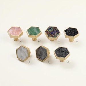 May include: Seven decorative cabinet knobs with hexagonal faces and brass bases. The knobs feature various designs, including pink, green, iridescent, black, white, gray, and black leather textures. Each knob is labeled with a letter from A to G.