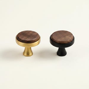 May include: Two cabinet knobs with a round, dark wood top. One knob has a gold-colored base and trim, while the other has a black base and trim. The knobs are designed for furniture and home decor.