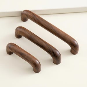 May include: Three wooden cabinet pulls in varying sizes. The pulls are made of dark brown wood with a curved handle design. The pulls are arranged diagonally, showcasing their different lengths.