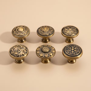 May include: Six antique brass cabinet knobs with various floral and scroll designs. The knobs have a round shape and are mounted on short stems. The finish is a warm, aged brass color, adding a vintage touch to any furniture piece.