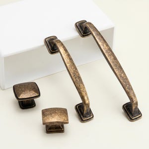 May include: A set of antique bronze cabinet hardware, comprising two handles and two square knobs. The hardware features a distressed finish, giving a vintage appearance. Ideal for updating kitchen or bathroom cabinets.