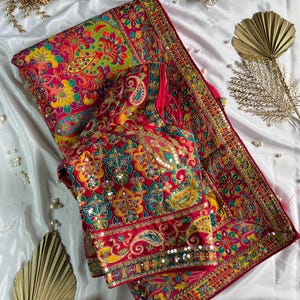 May include: A vibrant fuchsia saree and matching blouse set featuring intricate multi-coloured floral and paisley embroidery. The fabric is adorned with shimmering sequins and metallic thread work, showcasing a dense, ornate pattern throughout the garment.