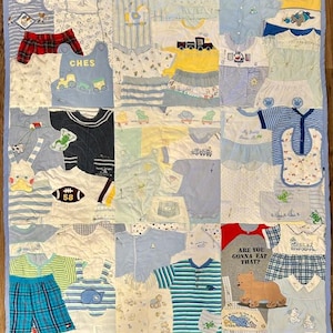 May include: A patchwork quilt made from baby clothes in various shades of blue, white, yellow, and red. The quilt features a variety of baby shirts, shorts, and bibs, some with printed designs and text. The quilt is bordered with a light blue fabric.