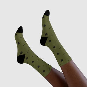 Può includere: Calzini verde oliva con tallone e punta neri. I calzini presentano un motivo ripetuto di piccole illustrazioni nere di tacos e linee. I calzini sono mostrati sulle gambe di una persona.
