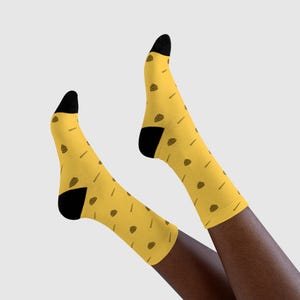 Può includere: Un paio di calzini gialli con talloni e punte neri. I calzini presentano un motivo con piccole illustrazioni marroni di tacos e patatine fritte. I calzini sono mostrati sulle gambe di una persona.