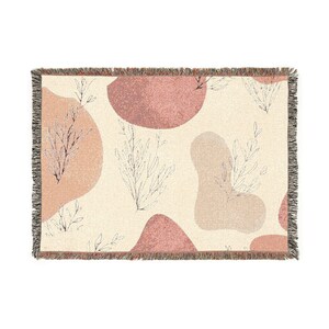 May include: A woven throw blanket with a cream background and a pattern of abstract shapes in shades of pink and beige. Delicate black line drawings of plants are scattered across the surface. The blanket has a fringed edge.