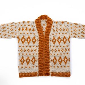 May include: Cream-colored crochet cardigan with a geometric pattern in shades of orange. The open-front sweater features a shawl collar and long sleeves. The crochet design includes diamond and zig-zag patterns.