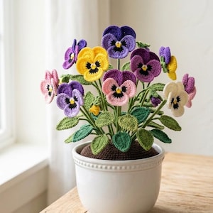 May include: A handcrafted crochet pansy flower arrangement in a white ceramic pot. The colorful flowers include purple, yellow, pink, and white petals with green leaves. The pot sits on a wooden table, creating a charming home decor piece.