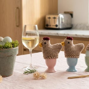 Könnte beinhalten: Ein Stillleben mit Ostermotiven. Ein Topf mit Eiern, ein Weinglas und drei gestrickte Hühner-Eierwärmer in rosa, blauen und grünen Eierbechern. Ein goldener Löffel und Trockenblumen ergänzen die Szene.