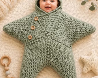 Puede incluir: Un saco de dormir para bebé en forma de estrella, tejido a ganchillo en color verde salvia, con capucha y tres botones de madera en el lateral. La prenda presenta un patrón de punto texturizado con líneas en relieve que irradian desde el centro.