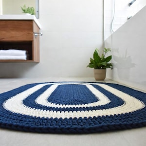 May include: Oval-shaped, navy blue and white crochet rug. The rug features a navy blue border with two concentric white stripes in the center. The rug is designed for a bathroom setting.