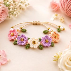 May include: A bracelet featuring crocheted flowers in shades of pink, lavender, cream, and peach, accented with small green crocheted leaves, gold-toned beads, and white pearls. The piece includes a beige cord with an adjustable sliding knot closure.
