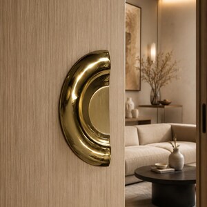 May include: A polished gold-toned semi-circular door handle mounted on a light-colored wood grain sliding door. The handle features a smooth, curved, recessed design with a reflective metallic finish, contrasting against the vertical wood texture.