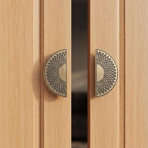 May include: Two semi-circular door handles made of antique brass with a detailed sunburst or petal-like cutout pattern. The handles are mounted on light-colored wooden double doors with vertical grain, positioned symmetrically on each door panel.