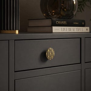 Puede incluir: Un cajón de mueble gris oscuro con un pomo circular de latón que presenta un patrón geométrico de arcos repetidos. Sobre el cajón, dos libros están apilados, uno titulado Chanel Catwalk y el otro Live Beautiful, junto a un jarrón de vidrio oscuro.
