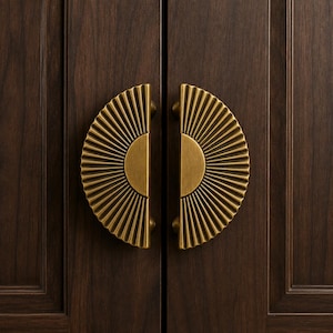 May include: Two semi-circular brass cabinet pulls with a sunburst pattern of radiating ridges. Each handle features a smooth central semi-circle flanked by textured, fanned-out lines, mounted vertically on dark brown wooden cabinet doors.