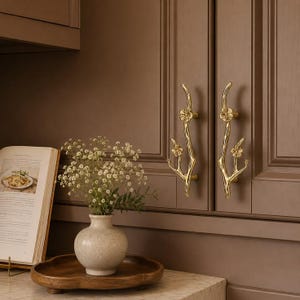 May include: Two gold-toned metal cabinet handles shaped like tree branches with small flower blossoms attached. The handles are mounted vertically on brown cabinet doors, featuring a polished finish and detailed floral accents on each branch.