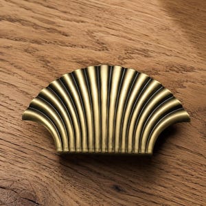 Op de afbeelding: Een messingkleurige meubelgreep in de vorm van een waaier of een schelp. Het beslag is voorzien van een reeks verticale, verhoogde ribbels die vanuit de basis naar de gebogen bovenrand uitstralen, wat een textuurrijke, halfronde waaier silhouet creëert.