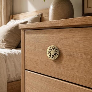 Puede incluir: Un tirador de mueble circular en tono latón con un diseño de sol recortado y patrones en forma de llama. El tirador está montado en la parte frontal de un cajón de madera clara, que forma parte de un mueble de mayor tamaño.