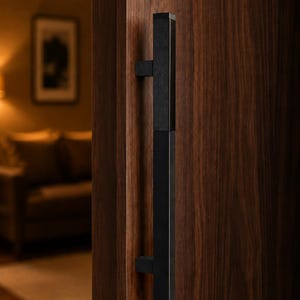May include: A long, vertical, matte black rectangular door handle mounted onto a dark brown wooden door. The handle features a clean, linear design with two square mounting brackets connecting the bar to the wood surface, creating a sharp contrast.