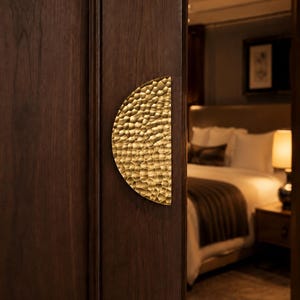May include: A semi-circular door handle with a hammered, textured gold finish mounted on a dark brown wooden door. The handle features a distinct dimpled surface pattern that catches the light, providing a tactile grip for opening the door.