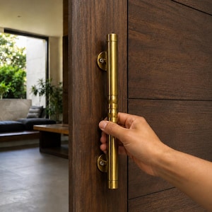 May include: A long, cylindrical, brushed brass door handle mounted vertically on a dark brown wooden door. The handle features a smooth, metallic finish with two circular mounting plates secured by visible screws at the top and bottom ends.