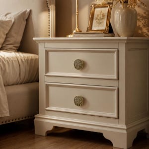 May include: A cream-colored wooden nightstand featuring two drawers with ornate, circular gold and black metal knobs. The nightstand has a rectangular frame with raised panel detailing on the drawer fronts and stands on four short, curved feet.