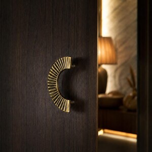Op de afbeelding: Een halfronde kastgreep van messing met een geplooide, waaiervormige textuur, gemonteerd op een donkerbruine houten deur. De greep heeft een metallic gouden afwerking en een gebogen ontwerp dat uitsteekt vanaf het vlakke oppervlak.