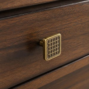 Könnte beinhalten: Ein quadratischer Messing-Möbelknopf mit Gittermuster auf der Vorderseite, befestigt an einer dunkelbraunen Holzschublade. Der Knopf hat eine metallische Oberfläche mit einer vertieften Mitte, die ein erhabenes Kreuzschraffur-Design aufweist.