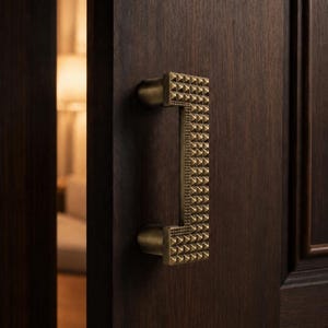 Puede incluir: Un tirador de puerta rectangular de latón con una textura de tachuelas piramidales montado en una puerta de madera de color marrón oscuro. El tirador presenta un diseño geométrico repetitivo en su superficie y se fija con dos soportes cilíndricos de latón.