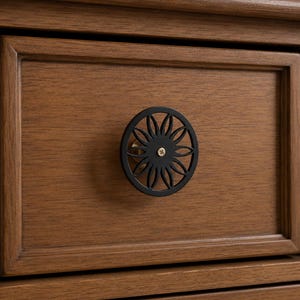 May include: A circular black metal cabinet knob with a cut-out floral or sunburst pattern, featuring a small gold-toned screw in the centre. The knob is mounted on a medium-brown wooden drawer front with a rectangular frame detail.