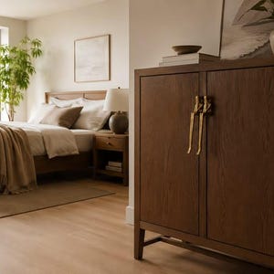 May include: A dark brown wooden cabinet with two doors and long, vertical gold-toned metal handles. The cabinet features a visible wood grain texture and stands on four short legs, positioned in a room with a bed and a nightstand in the background.