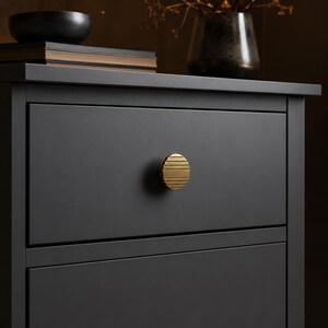 May include: A matte black drawer front featuring a single circular brass knob. The knob has a textured surface with horizontal grooves. The drawer is part of a larger piece of furniture with a flat top surface and a clean, solid color finish.