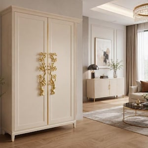 May include: A tall, off-white wardrobe featuring two doors with long, vertical gold-colored handles shaped like flowering branches. The cabinet has a smooth, matte finish, a rectangular frame design on each door, and stands on four short, tapered legs.